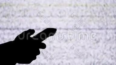 一只手拿着电视遥控器在电视屏幕背景上的剪影，带有白色的静态噪音